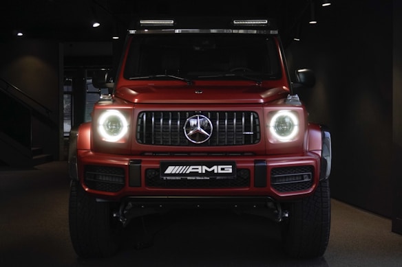 A red, powerful SUV with distinctive front headlights is displayed in a dimly lit showroom setting. The vehicle features a prominent grille with a recognizable emblem at the center and has a sleek, modern design that underscores its luxury and performance attributes.