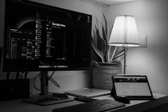 A monochrome workspace featuring a large monitor displaying a music playlist titled 'late night vibes' and a calendar for December 2023. A tablet sits on a desk displaying a webpage alongside a wireless keyboard and mouse. A lamp with a pleated shade is lit, casting soft lighting over the scene. A potted plant is placed behind the lamp, contributing to a cozy ambiance.