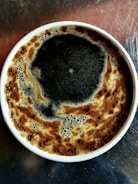 A cup of frothy coffee with a rich layer of dark crema on top, set against a metallic surface.