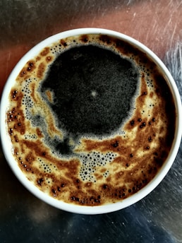A cup of frothy coffee with a rich layer of dark crema on top, set against a metallic surface.