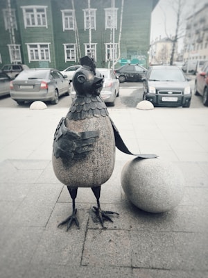A whimsical sculpture of a bird stands on a sidewalk, crafted with a combination of metal and stone, featuring intricate details on its head and feathers. The bird is accompanied by a large spherical object. Behind the sculpture, several cars are parked along the street in front of a green building with white-framed windows.