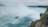 A large, powerful waterfall cascades down a steep cliff, creating a misty atmosphere. A boat with tourists onboard navigates the turbulent waters near the base of the waterfall. The surrounding landscape is lush and green, with rocky outcrops and trees bordering the scene.