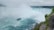 A large, powerful waterfall cascades down a steep cliff, creating a misty atmosphere. A boat with tourists onboard navigates the turbulent waters near the base of the waterfall. The surrounding landscape is lush and green, with rocky outcrops and trees bordering the scene.