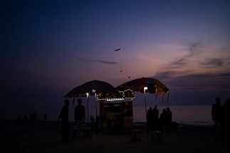 A colorful food van parked by the beach at sunset with people enjoying carnival snacks nearby.