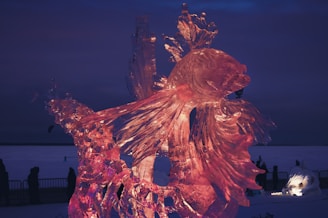 A large, intricately detailed ice sculpture of a fish with flowing fins is prominently displayed. The sculpture is illuminated with pink lights, creating a warm glow against a dark, nighttime background. Silhouettes of people and barriers can be seen in the distance, suggesting an outdoor event.