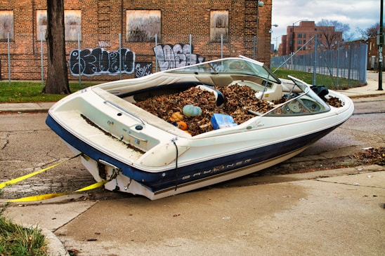 A small motorboat is parked on a city street, filled with fallen leaves and some debris. The boat is not in water, but rather on a pavement next to an old brick building with graffiti. The setting appears urban and somewhat neglected, with bare tree trunks, a chain-link fence, and cloudy skies.