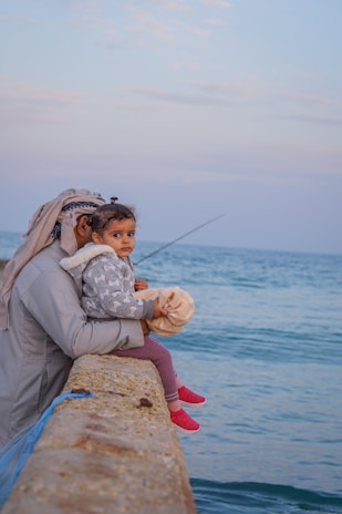 A man is sitting by the sea with a young child on his lap. He is holding a fishing rod while the child is looking towards the camera. The sky is partly cloudy with a pale blue hue. Both the man and the child are dressed warmly, indicating a cooler climate.