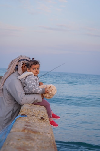 A man is sitting by the sea with a young child on his lap. He is holding a fishing rod while the child is looking towards the camera. The sky is partly cloudy with a pale blue hue. Both the man and the child are dressed warmly, indicating a cooler climate.