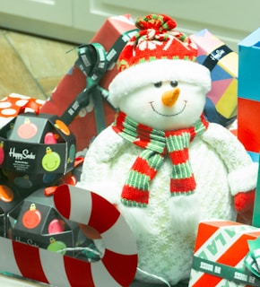 A cheerful snowman figurine wearing a red and white knit hat and a striped scarf is surrounded by a collection of colorful, wrapped gift boxes and a candy cane decoration. The gifts include a variety of festive patterns and colors, with a visible 'Happy Socks' label suggesting sock-themed gifts.