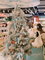 A Christmas tree decorated with blue and silver ribbons, plush toys, and other ornaments stands in the foreground. Behind it, there is a store with shelves filled with stuffed animals and toys. A person is interacting with a large stuffed dog placed in a bin with the 'Jellycat London' logo.