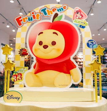 A colorful display featuring a character with a smiling face and an apple-themed design, surrounded by various decorations. The background includes small decorative elements and stars on stands. The design incorporates playful elements with bright colors, drawing attention to its cheerful theme in a retail environment.