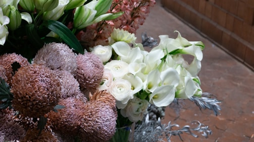 A vibrant arrangement of flowers including white calla lilies, roses, and various textured blooms in shades of pink and cream. Lush green foliage frames the flowers, creating a rich display. The background features a subtle, neutral-toned floor with a tile pattern.