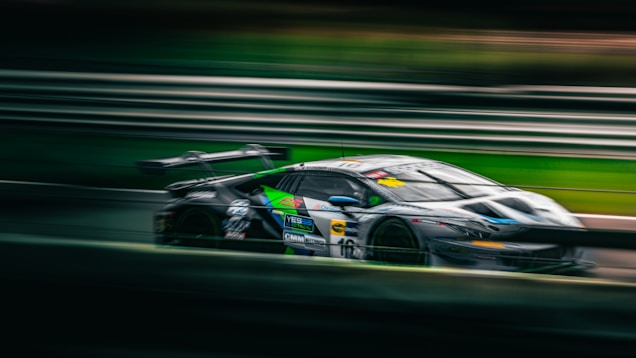 A sleek, high-performance race car speeds along a racetrack, creating a motion blur effect that emphasizes its velocity. The car features a combination of dark colors, including black and green, with various sponsor logos visible on its body.