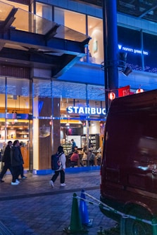 A photo of an evening street scene with people walking past a well-lit Starbucks caf&eacute;. The caf&eacute; has large glass windows showing customers inside. Above the Starbucks, there is a sign for 'nana's green tea'. There are also blue lights illuminating parts of the building. A red parked van and some green cones are visible in the foreground.
