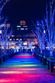 A nighttime urban scene featuring a pathway illuminated by colorful lights in a gradient pattern from red to blue. Trees lining the path are adorned with bright blue string lights, and a group of people is gathered in the distance. Buildings with illuminated signs are visible in the background.