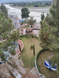 An aerial view of a riverside area with a road running alongside the river. Several trees are scattered around the landscape, including a large tree near the riverbank. A small building with a rooftop seating area is surrounded by green lawns and pathways. On the right side, there is a blue-and-white outdoor umbrella providing shade over patio furniture.