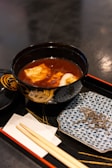 A bowl of soup with tofu and other ingredients is placed on a black lacquer tray. The bowl is ornately decorated and accompanied by a small plate with a patterned design. Wooden chopsticks rest on a white paper napkin placed on the tray.