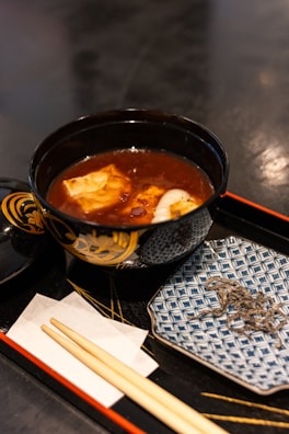 A bowl of soup with tofu and other ingredients is placed on a black lacquer tray. The bowl is ornately decorated and accompanied by a small plate with a patterned design. Wooden chopsticks rest on a white paper napkin placed on the tray.