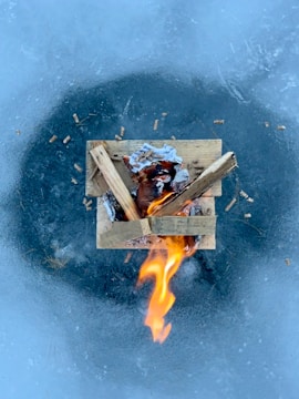A small fire burns on top of an icy surface, surrounded by wooden planks and remnants of paper. The flame is vivid against the cold blue of the ice.