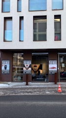 A modern building with multiple rectangular windows above a gallery entrance. The gallery features posters advertising a Tyler Shields exhibition, with artworks displayed inside. A traffic cone is situated on the snowy sidewalk in front of the gallery.