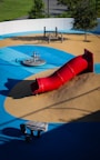 A playground featuring a vibrant red tube slide on a mound of artificial turf surrounded by blue and tan padded surfaces. The area includes a spinning seat and a climbing play structure made of wood. Trees and greenery are visible around the perimeter.