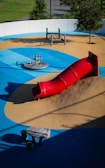 A playground featuring a vibrant red tube slide on a mound of artificial turf surrounded by blue and tan padded surfaces. The area includes a spinning seat and a climbing play structure made of wood. Trees and greenery are visible around the perimeter.