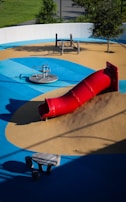 A playground featuring a vibrant red tube slide on a mound of artificial turf surrounded by blue and tan padded surfaces. The area includes a spinning seat and a climbing play structure made of wood. Trees and greenery are visible around the perimeter.