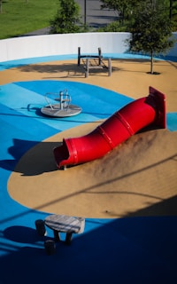 A playground featuring a vibrant red tube slide on a mound of artificial turf surrounded by blue and tan padded surfaces. The area includes a spinning seat and a climbing play structure made of wood. Trees and greenery are visible around the perimeter.