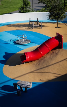 A playground featuring a vibrant red tube slide on a mound of artificial turf surrounded by blue and tan padded surfaces. The area includes a spinning seat and a climbing play structure made of wood. Trees and greenery are visible around the perimeter.