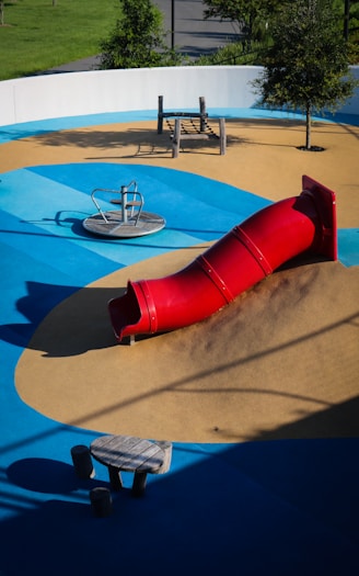 A playground featuring a vibrant red tube slide on a mound of artificial turf surrounded by blue and tan padded surfaces. The area includes a spinning seat and a climbing play structure made of wood. Trees and greenery are visible around the perimeter.