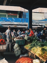 A bustling outdoor market with vendors and customers interacting. Various fresh vegetables such as leafy greens and cabbages are displayed on the stalls. People are seen walking around, some wearing helmets. The market is partially covered, and the background shows a blue wall and a building with several metal shutters.