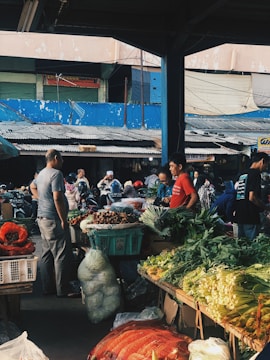 A bustling outdoor market with vendors and customers interacting. Various fresh vegetables such as leafy greens and cabbages are displayed on the stalls. People are seen walking around, some wearing helmets. The market is partially covered, and the background shows a blue wall and a building with several metal shutters.
