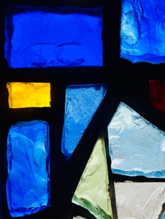 Several pieces of colorful stained glass are arranged in a pattern. The fragments include various shapes such as rectangles and triangles, with a mix of textures. The colors range from deep blue and bright yellow to shades of green and light blue.