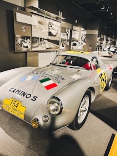 A vintage silver race car with racing decals and a notable number 154 on a yellow plate at the front. The car is displayed in a museum setting with historical racing photographs and memorabilia on the walls. The vehicle features prominent headlamps and a sleek, aerodynamic design.