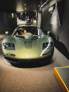 A sleek green sports car is prominently displayed in an indoor showroom. The vehicle's aerodynamic design and luxurious interior can be seen clearly. Overhead lighting accentuates the car's glossy finish, and a photograph of a road is mounted on the wall nearby.