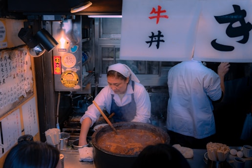 A woman wearing a white apron is cooking in a small kitchen. She is stirring a large pot filled with a brown stew or sauce. To her right, there's another person wearing a white shirt facing away. Various kitchen utensils and ingredients such as chopsticks and eggs are visible on the counter. The walls are adorned with Japanese writing and stickers. Overhead, a sign with Japanese characters hangs.