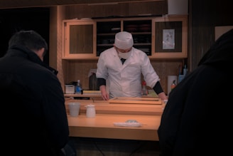A chef dressed in a white uniform and hat works behind a wooden counter. He appears to be preparing food, possibly in a sushi restaurant. There are two patrons seated at the counter with dark coats. Various condiments and utensils are visible on the counter. Wooden shelves and a wall with traditional decor form the background.