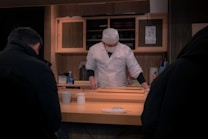 A chef dressed in a white uniform and hat works behind a wooden counter. He appears to be preparing food, possibly in a sushi restaurant. There are two patrons seated at the counter with dark coats. Various condiments and utensils are visible on the counter. Wooden shelves and a wall with traditional decor form the background.