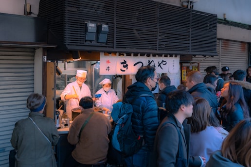 A busy street food stall with people gathered around, some waiting in line and others being served. Two chefs in white attire and face masks are preparing food behind the counter. The stall is set up in an urban environment with a sign above it displaying Japanese characters.