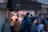 A busy street food stall with people gathered around, some waiting in line and others being served. Two chefs in white attire and face masks are preparing food behind the counter. The stall is set up in an urban environment with a sign above it displaying Japanese characters.