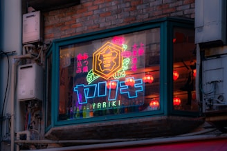 A neon sign is displayed in a window, surrounded by a brick wall. The sign is vibrant with colors, including blue, pink, and yellow, and features Japanese characters. Inside the window, red paper lanterns can be seen, adding a warm glow to the interior.