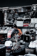 A cluttered market stall is filled with various goods, including clothes, shoes, and packaged items. A person is sitting among the items, appearing to organize or sort through them. Surrounding shelves are densely packed with folded shirts and plastic packages. There are signs with messages such as 'Don't Touch' in both English and another language.