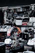 A cluttered market stall is filled with various goods, including clothes, shoes, and packaged items. A person is sitting among the items, appearing to organize or sort through them. Surrounding shelves are densely packed with folded shirts and plastic packages. There are signs with messages such as 'Don't Touch' in both English and another language.