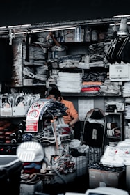 A cluttered market stall is filled with various goods, including clothes, shoes, and packaged items. A person is sitting among the items, appearing to organize or sort through them. Surrounding shelves are densely packed with folded shirts and plastic packages. There are signs with messages such as 'Don't Touch' in both English and another language.