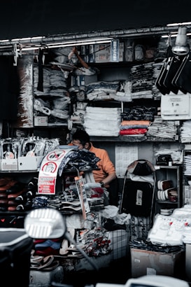 A cluttered market stall is filled with various goods, including clothes, shoes, and packaged items. A person is sitting among the items, appearing to organize or sort through them. Surrounding shelves are densely packed with folded shirts and plastic packages. There are signs with messages such as 'Don't Touch' in both English and another language.