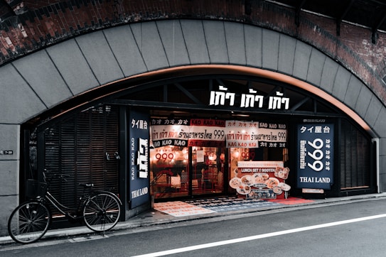 A storefront with a distinctly arched entrance, featuring numerous signs in a foreign language displayed prominently. The interior appears to be warmly lit with visible hanging lights. A bicycle is parked outside on the street against the wall, adding an urban feel. The text suggests this may be a restaurant or food establishment with promotional offers.