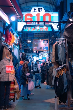 A bustling shopping arcade illuminated with bright neon signs. Various clothing items are displayed on racks lining the narrow walkway. A person in a blue jacket and skirt stands by a sale sign, while several other people mill about, browsing and chatting.