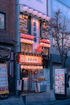 An inviting street view of a small Japanese eatery adorned with bright paper lanterns and an array of signs in Japanese script. The entrance is warmly lit with a mix of red and white lights, and there is a prominent Daruma doll decoration. Menus and promotional posters flank the doorway while a tree with autumn leaves stands nearby.