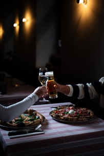 Two people are clinking their drinks, one holding a glass of wine and the other a glass of beer, while seated at a table with two pizzas. The setting appears to be a cozy, dimly-lit restaurant with warm lighting from wall-mounted lamps.