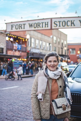 A woman stands on a cobblestone street in front of a large sign that reads 'Fort Worth Stock Yards'. She is wearing a quilted jacket, a scarf, and is carrying a purse. Behind her, a bustling street scene unfolds with people walking and shops illuminated with lights.